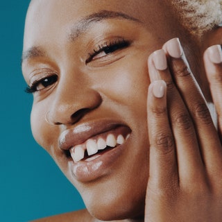 woman with blonde buzz cut putting on skin-care product while smiling against a teal background